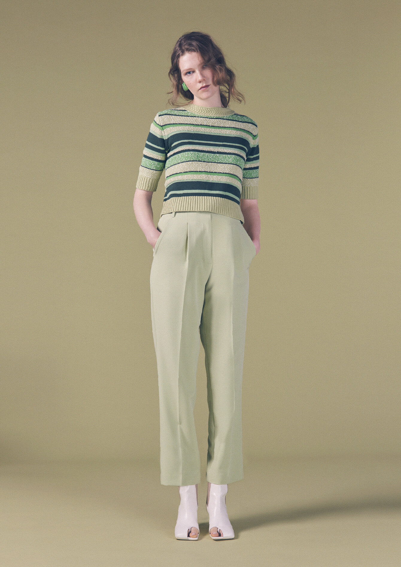 緑横線のトップスと薄い緑のパンツと白い靴を入ってズボンのポケットに両手を入れて前を見て立っている女性モデルの画像