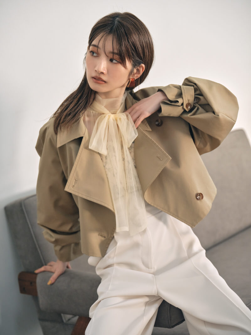 ベージュのジャケットを着てソファの手すりに座って斜め右を見ている女性モデルの画像