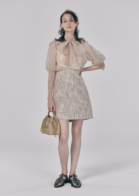 ベージュ色のトップスとスカートでカバンを持って立っている女性モデルの画像