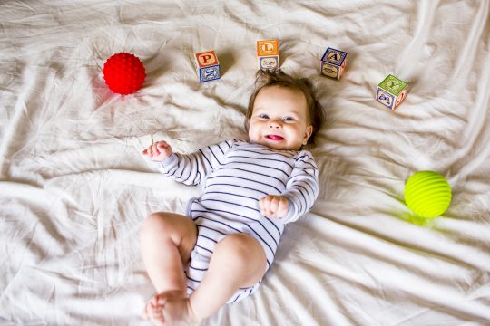 A newborn baby playing with blocks and balls in the bedroom of a home next to a window.